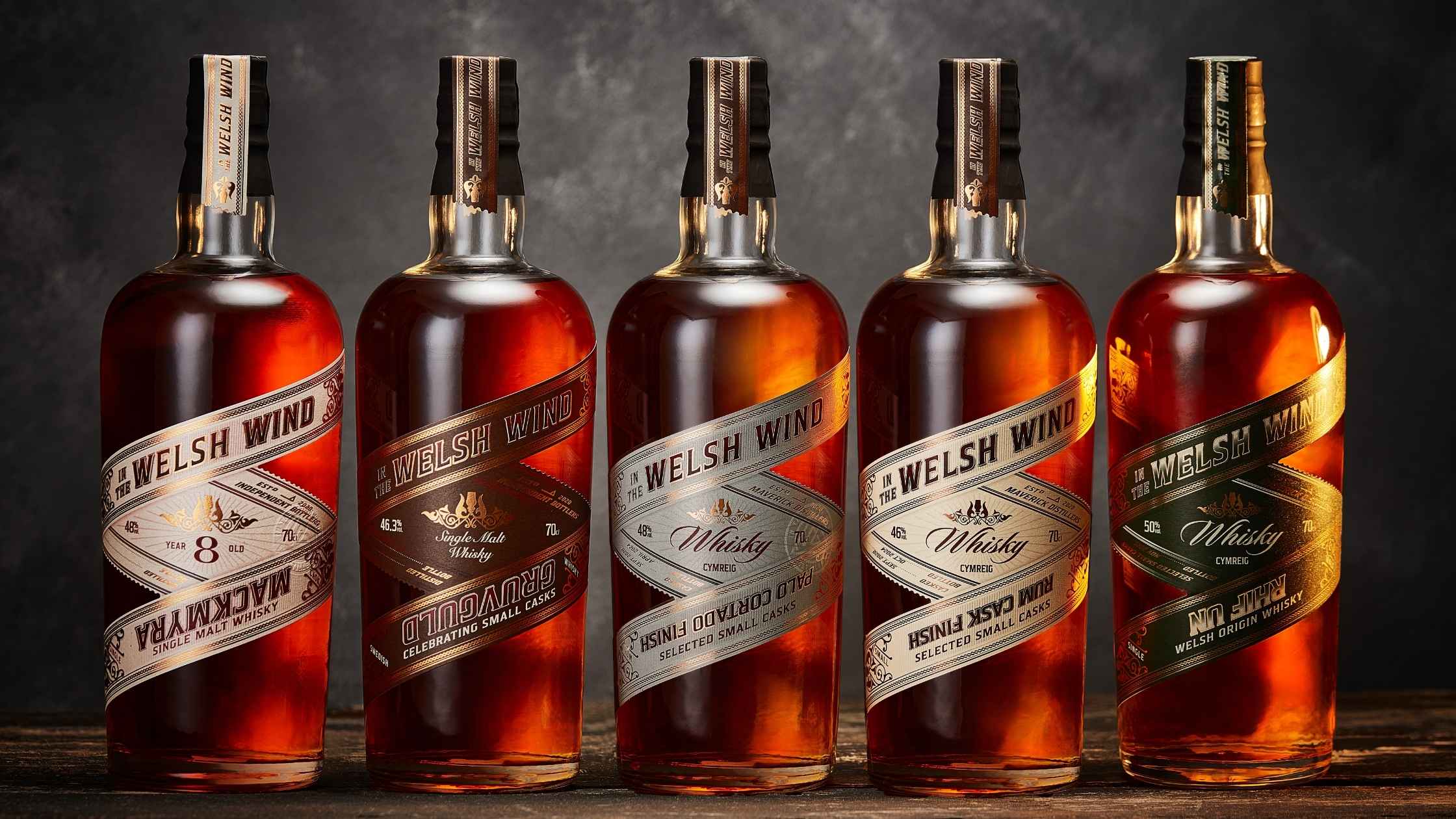 A row of five In the Welsh Wind Welsh whisky bottles on a wooden table against a dark background.