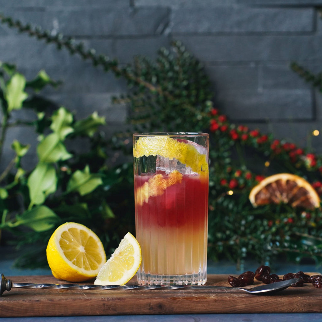 tall cocktail glass with a two tone pink and yellow cocktail against a festive background of holly and berries