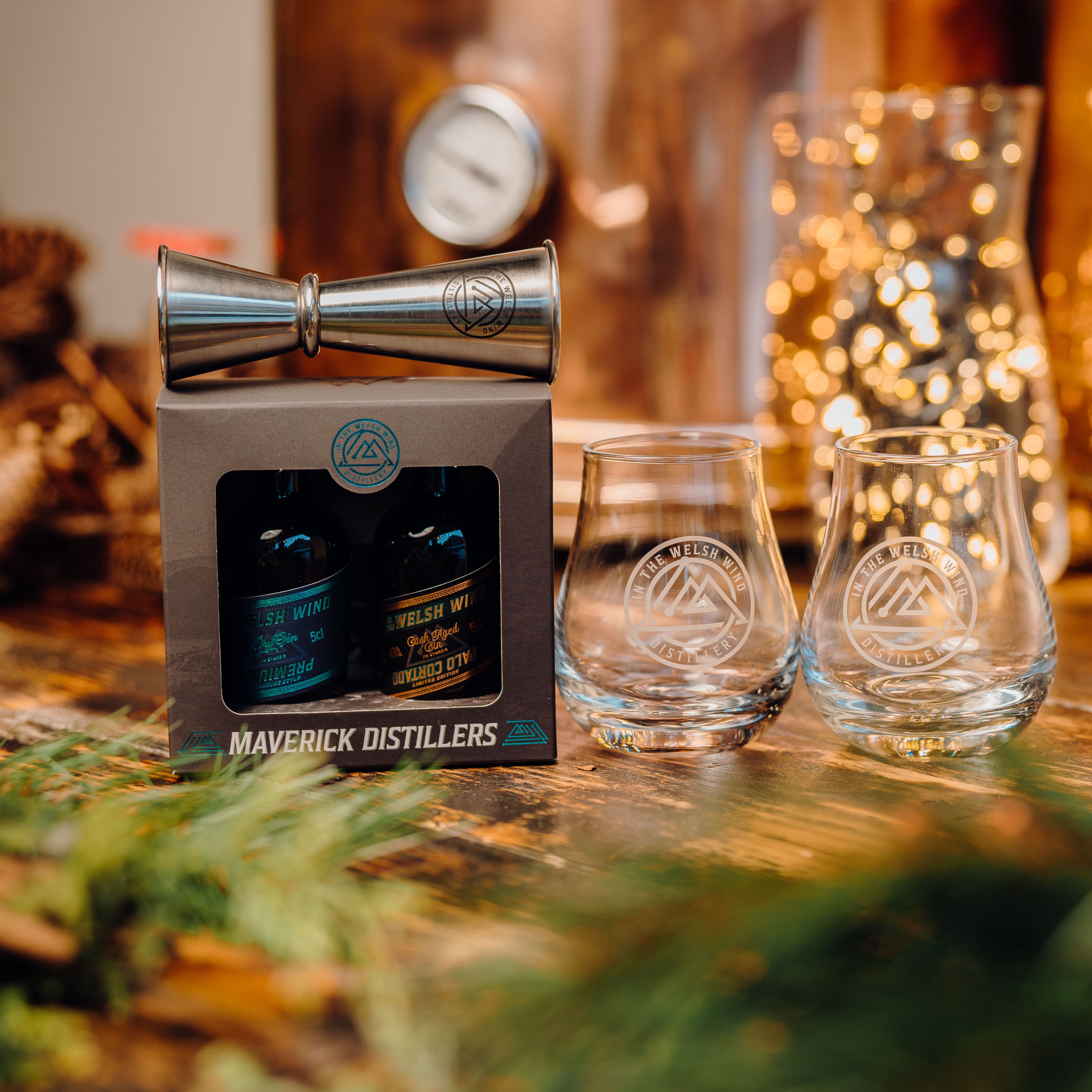 A box of gin miniatures with a pair of engraved glasses and a double ended measure from In the Welsh Wind Welsh gin distillery against a copper background