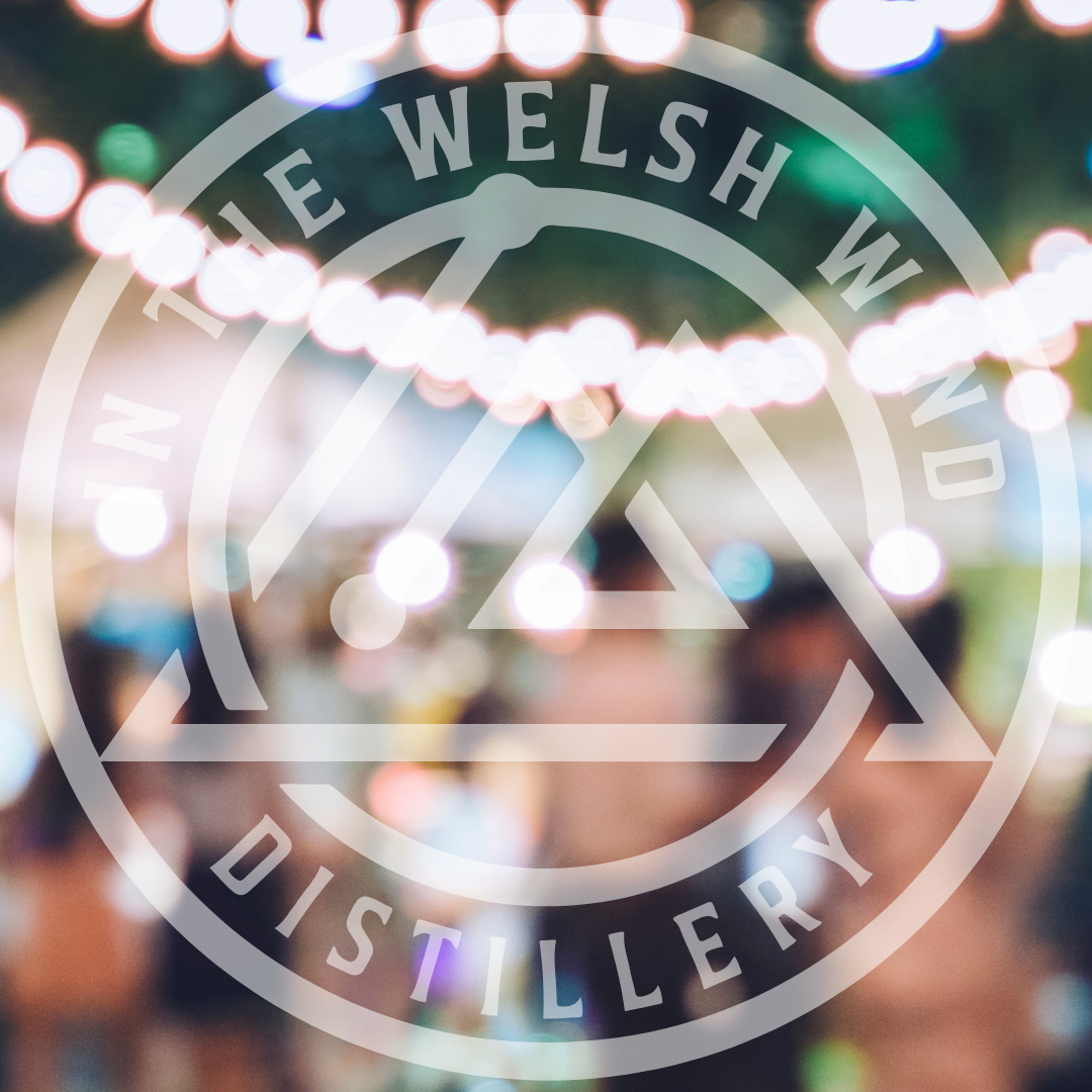 Strings of warm festoon lights glowing over an evening crowd, with the In the Welsh Wind Distillery logo